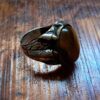 A silver-toned metal ring featuring an oval-shaped, smooth, brown tiger's eye gemstone set in the center. The band of the ring displays intricate, raised leaf-like engravings and textured detailing along the sides of the metal setting. — Khodamdjin