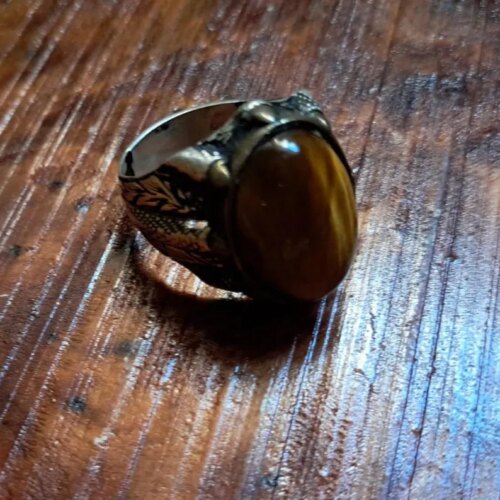 A silver-toned metal ring featuring an oval-shaped, brown and gold tiger's eye gemstone set in a pronged mounting. The band of the ring displays intricate, engraved leaf-like patterns and a darkened, oxidized finish. — Khodamdjin