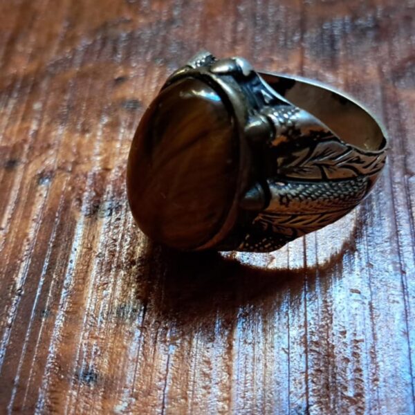 A silver-toned metal ring featuring a large, oval-shaped, brown tiger's eye gemstone set in the center. The band of the ring is decorated with intricate, engraved leaf-like patterns and textured detailing along the sides. — Khodamdjin