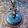 A circular pendant featuring a smooth, light blue cabochon stone set within a textured, silver-toned metal frame that resembles coiled snake scales. A detailed, three-dimensional silver-toned snake head rests atop the stone. — Khodamdjin