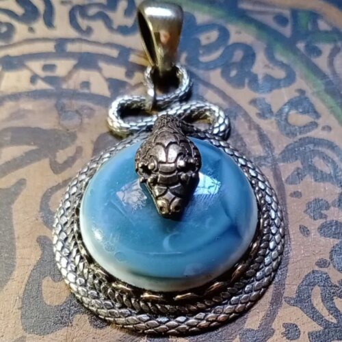 A circular pendant featuring a smooth, light blue cabochon stone set within a textured, silver-toned metal frame that resembles coiled snake scales. A detailed, three-dimensional silver-toned snake head rests atop the stone. — Khodamdjin