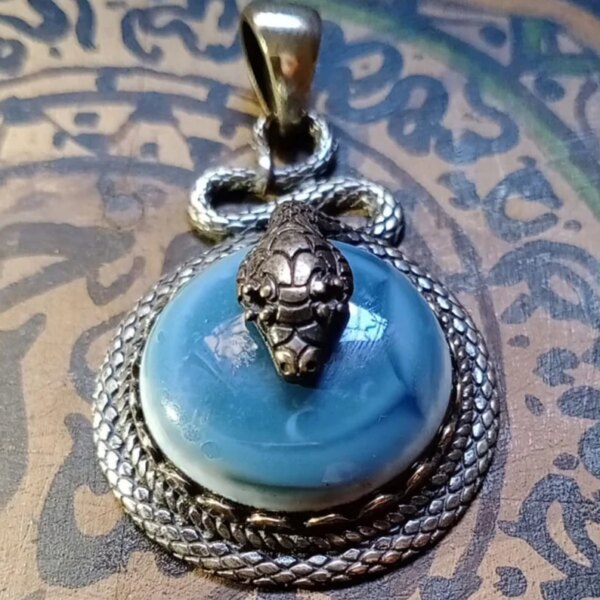 A circular pendant featuring a smooth, light blue cabochon stone set within a textured, silver-toned metal frame that resembles coiled snake scales. A detailed, three-dimensional silver-toned snake head rests atop the stone. — Khodamdjin