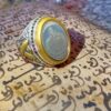 A silver and gold-toned ring featuring an oval-shaped center stone with an engraved Arabic script design. The band displays intricate metalwork patterns on the sides, surrounding the central stone set within a smooth gold-colored bezel. — Khodamdjin