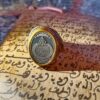 A gold-toned ring featuring an oval face with a dark gray inlay. The center of the inlay displays intricate silver-colored Arabic calligraphy. The band of the ring has detailed engravings along the sides, providing a textured appearance. — Khodamdjin