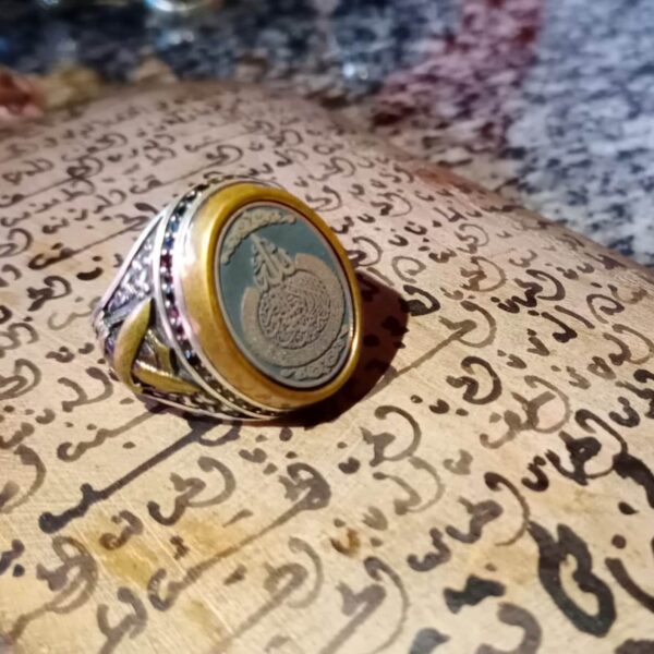 A silver ring featuring a circular gold-toned bezel surrounding a dark center inlay with intricate Arabic calligraphy. The band displays detailed metalwork with small embedded stones along the sides and a polished finish. — Khodamdjin