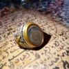 A silver and gold-toned ring featuring an oval-shaped dark center stone engraved with intricate Arabic calligraphy. The metal band displays detailed relief patterns along the sides, providing a textured appearance to the jewelry piece. — Khodamdjin