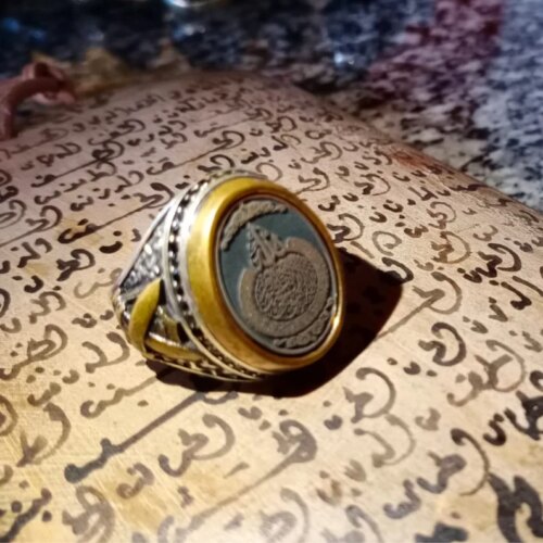 A silver and gold-toned ring featuring an oval-shaped dark center stone engraved with intricate Arabic calligraphy. The metal band displays detailed relief patterns along the sides, providing a textured appearance to the jewelry piece. — Khodamdjin