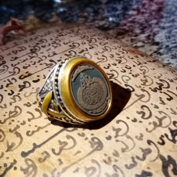 A silver and gold-toned ring featuring an oval-shaped dark center stone engraved with intricate Arabic calligraphy. The metal band displays detailed relief patterns along the sides, providing a textured appearance to the jewelry piece. — Khodamdjin