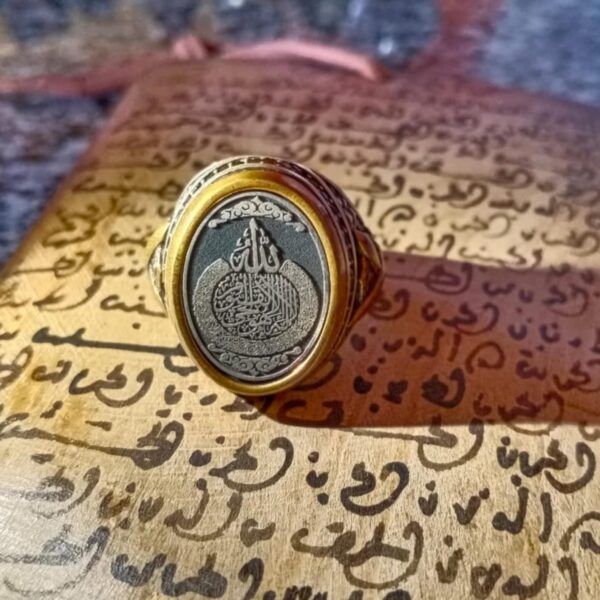 A gold-toned ring featuring an oval face with a dark gray inlay. The center of the inlay displays intricate silver-colored Arabic calligraphy. The band of the ring has detailed engravings along the sides, providing a textured appearance. — Khodamdjin