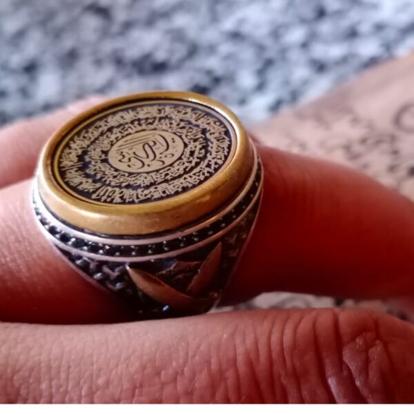 A silver-toned ring featuring a circular top with a gold-colored rim and a black center engraved with intricate Arabic calligraphy. The sides of the ring band display a textured, dark metal finish with a subtle gold-toned accent. — Khodamdjin