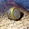 A silver-toned metal ring featuring an ornate, textured band with intricate cut-out details. The circular face of the ring is framed by a smooth, mustard-yellow border and contains a central medallion with engraved Arabic script. — Khodamdjin