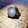 A silver and gold-toned ring featuring an oval black stone inlay. The stone is engraved with intricate white Arabic calligraphy. The metal band has a detailed, textured design along the sides, providing a contrast to the smooth, polished bezel surrounding the central stone. — Khodamdjin