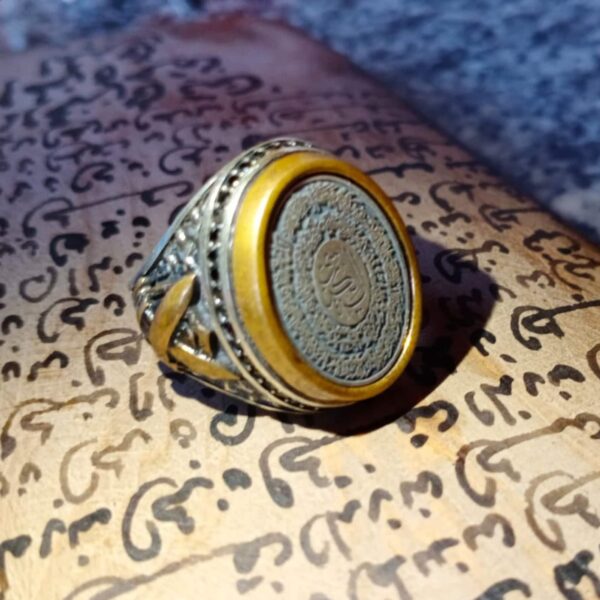 A silver-toned metal ring featuring an ornate, textured band with intricate cut-out details. The circular face of the ring is framed by a smooth, mustard-yellow border and contains a central medallion with engraved Arabic script. — Khodamdjin