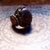A silver-toned ring featuring a large, oval-shaped dark purple stone set in a decorative metal frame. The band is adorned with intricate gold-colored filigree patterns and small, circular turquoise-colored accents along the sides. — Khodamdjin