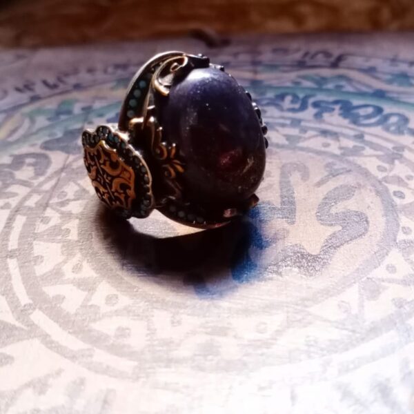 A silver-toned ring featuring a large, oval-shaped dark purple stone set in a decorative metal frame. The band is adorned with intricate gold-colored filigree patterns and small, circular turquoise-colored accents along the sides. — Khodamdjin