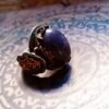 A silver ring featuring a large, oval, dark purple cabochon stone with subtle glitter. The metal band is decorated with intricate gold-toned scrollwork and small, embedded turquoise-colored stones along the sides and edges. — Khodamdjin