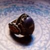 A silver ring featuring a large, oval, dark purple cabochon stone with gold-toned filigree accents and small black stones embedded along the sides. The metal band displays intricate engraved patterns and a dark, oxidized finish. — Khodamdjin