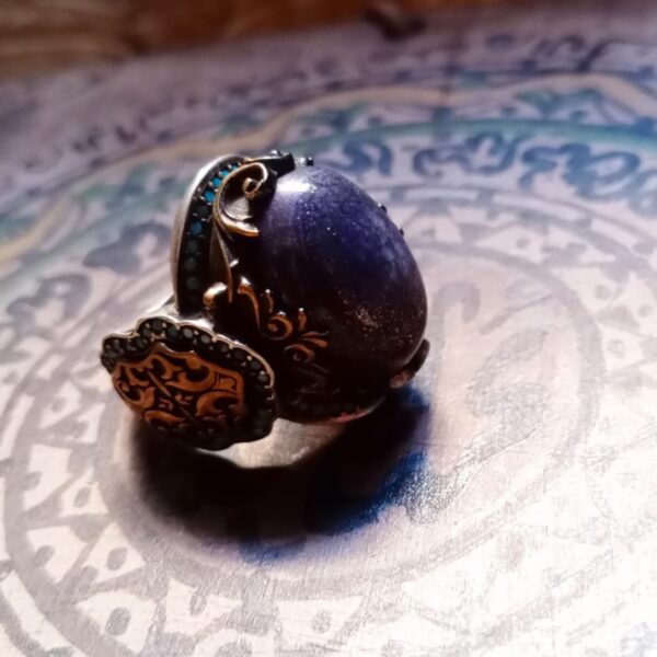 A silver ring featuring a large, oval, dark purple cabochon stone with subtle glitter. The metal band is decorated with intricate gold-toned scrollwork and small, embedded turquoise-colored stones along the sides and edges. — Khodamdjin