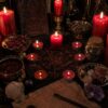 A black altar cloth featuring a red circular occult diagram with a central star, surrounded by several lit red pillar and tea light candles, bowls of dried herbs, a glass jar with cork stopper, crystals, and a piece of aged parchment. — Khodamdjin