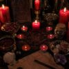 A black altar cloth featuring a red circular occult diagram with a central star, surrounded by red pillar and tea light candles, bowls of dried herbs, a glass jar of stones, a piece of parchment with handwritten text, and crystals. — Khodamdjin