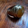A silver ring featuring an oval-shaped, translucent amber-colored stone set in a polished bezel. The band is crafted with intricate, dark-toned scrollwork engravings along the sides and includes a visible 925 hallmark stamp. — Khodamdjin