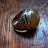 A silver ring featuring an oval-shaped, translucent amber-colored stone set in a band with intricate, dark, etched scrollwork patterns along the sides. The metal band has a polished finish and a visible hallmark stamp on the inner surface. — Khodamdjin