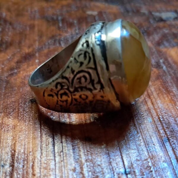 A silver ring featuring an intricate, dark-etched scrollwork pattern along the band and a large, smooth, amber-colored gemstone set in a polished silver bezel. The interior of the band is clearly stamped with the number 925. — Khodamdjin