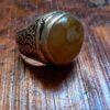 A silver ring featuring a large, round, polished amber-colored stone with white inclusions. The metal band is decorated with intricate, dark, recessed scrollwork patterns that wrap around the sides of the ring setting. — Khodamdjin