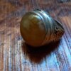 A silver ring featuring a large, smooth, round, amber-colored stone set in a metal band. The band is decorated with intricate, dark, engraved scrollwork patterns that wrap around the sides of the ring's setting. — Khodamdjin