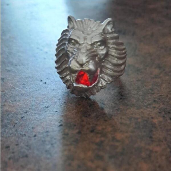 A silver-toned metal ring featuring a sculpted lion head design with a prominent mane. The lion's open mouth contains a single, faceted, bright red gemstone set within the center, creating a striking contrast against the metallic finish. — Khodamdjin