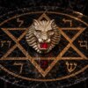 A metallic lion head ring featuring a detailed mane and a bright red gemstone set within its open mouth. The ring is positioned on a circular black marble surface decorated with a gold-colored hexagram and Hebrew characters. — Khodamdjin