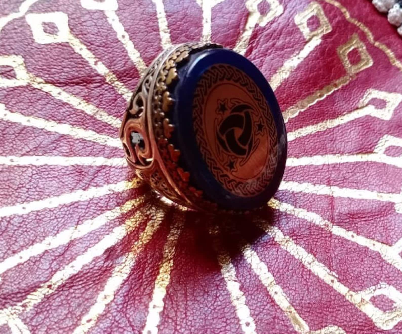 A gold-toned ring with an oval, dark blue stone. The stone features a gold-colored design with a central knot and stars. The ring has intricate filigree detailing on the sides and a gold band. — Khodamdjin