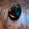 A silver-toned ring featuring an oval-shaped, faceted teal-colored stone set within an ornate, darkened metal frame. The band and setting display intricate, engraved patterns and a textured border surrounding the central gemstone. — Khodamdjin