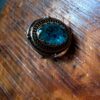 A ring featuring a large, oval-shaped, faceted blue stone set within a dark, textured metal frame. The setting includes intricate, darkened metalwork surrounding the central blue gemstone, creating a bold, detailed appearance. — Khodamdjin