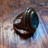 A metal ring featuring an oval-shaped, translucent light blue stone set within a dark, ornate frame. The band and setting display intricate, raised filigree patterns and a darkened, aged finish throughout the metal surface. — Khodamdjin