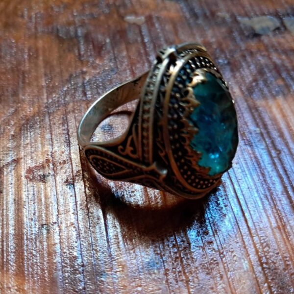 A metal ring featuring an oval-shaped, translucent light blue stone set within a dark, ornate frame. The band and setting display intricate, raised filigree patterns and a darkened, aged finish throughout the metal surface. — Khodamdjin