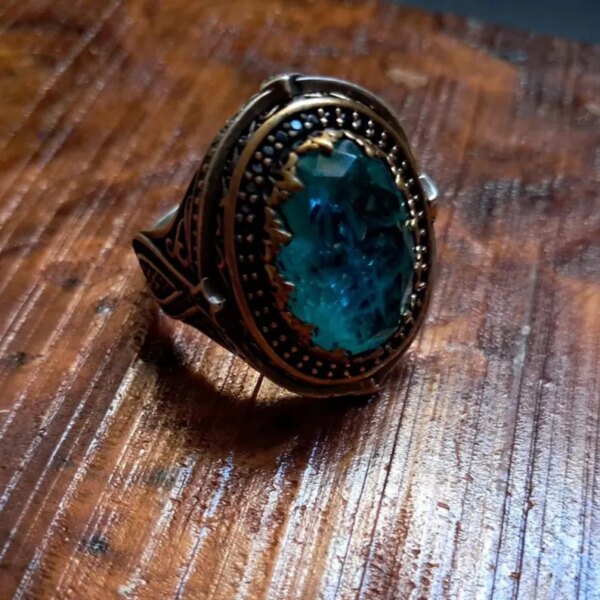 A silver-toned ring featuring an oval-shaped, faceted teal-blue stone set within a dark, ornate metal frame. The band and setting display intricate, engraved patterns and a textured, darkened finish surrounding the central gem. — Khodamdjin