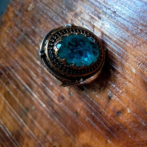 A ring featuring a large, oval-shaped, faceted blue stone set within a dark, textured metal frame. The setting includes intricate, darkened metalwork surrounding the central blue gemstone, creating a bold, detailed appearance. — Khodamdjin