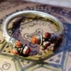A metallic cuff bracelet featuring a polished silver-tone band with engraved patterns. The open ends are adorned with decorative silver-tone caps, each set with three oval-shaped, shimmering copper-colored goldstone cabochons. — Khodamdjin