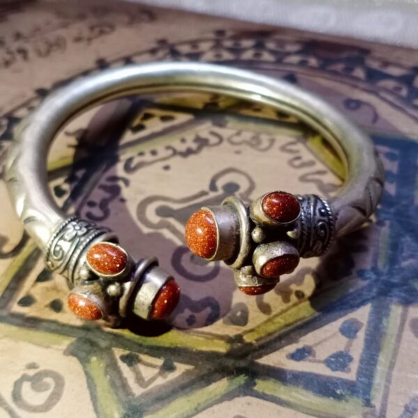 A metallic cuff bracelet featuring a polished silver-tone band with engraved patterns. The open ends are adorned with decorative silver-tone caps, each set with three oval-shaped, shimmering copper-colored goldstone cabochons. — Khodamdjin