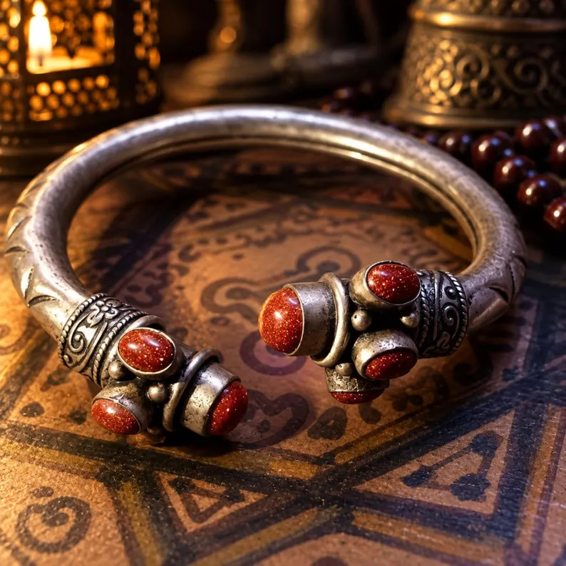 A silver-toned metal cuff bracelet featuring an open design with engraved patterns along the band. The two ends are decorated with multiple oval-shaped, reddish-brown glittery stones set in metal casings, creating a symmetrical appearance. — Khodamdjin