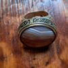 A gold-toned ring featuring a large, oval-shaped, banded agate stone with horizontal layers of white, tan, and brown. The metal band is wide and decorated with engraved, dark-filled symbols and characters around the setting. — Khodamdjin