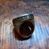 A metallic ring featuring an engraved band with Arabic calligraphy text that reads "Qul huwallahu ahad". The ring has a dark, oxidized finish in the recessed areas of the script and decorative patterns on the sides of the band. — Khodamdjin