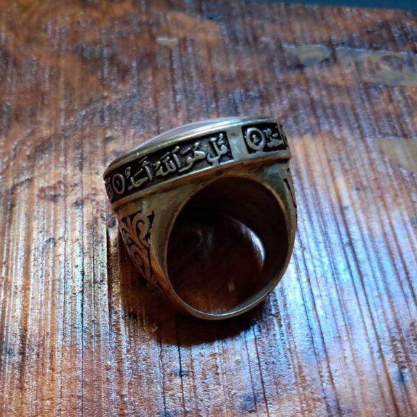 A metallic ring featuring an engraved band with Arabic calligraphy text that reads "Qul huwallahu ahad". The ring has a dark, oxidized finish in the recessed areas of the script and decorative patterns on the sides of the band. — Khodamdjin
