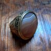 A silver-toned ring featuring a large, round, polished agate stone with visible brown and white horizontal banding. The metal band is decorated with intricate engraved scrollwork and a series of small circular symbols. — Khodamdjin