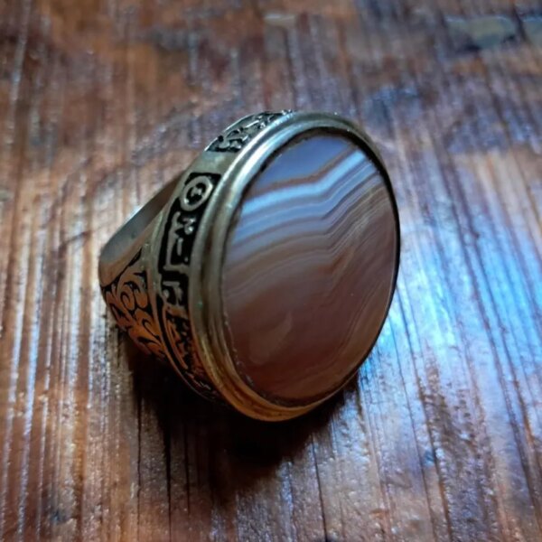 A silver-toned ring featuring a large, round, polished agate stone with visible brown and white horizontal banding. The metal band is decorated with intricate engraved scrollwork and a series of small circular symbols. — Khodamdjin