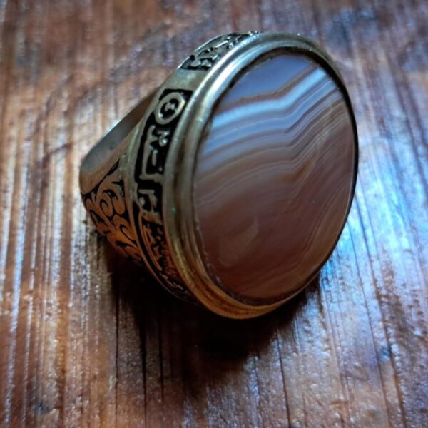 A silver-toned ring featuring a large, round, polished agate stone with horizontal bands of brown, white, and tan. The metal band is decorated with intricate black-filled engravings, including geometric patterns and symbols. — Khodamdjin