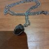A silver-toned metal chain necklace featuring a dark, textured, irregular-shaped stone pendant secured within a thin silver-colored wire wrap. The chain consists of elongated oval links connected by smaller circular links in a classic pattern. — Khodamdjin