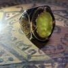 A silver ring with a large, oval, light-yellow gemstone. The ring has intricate gold-colored filigree and a dark, textured band. The gemstone is surrounded by a gold-colored rope-like border. — Khodamdjin