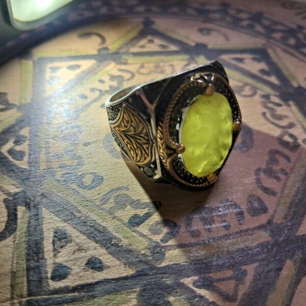 A silver ring with a large, light yellow gemstone. The ring has a black and gold filigree design on the sides and around the gemstone. The gemstone is oval-shaped and set in a gold bezel. — Khodamdjin